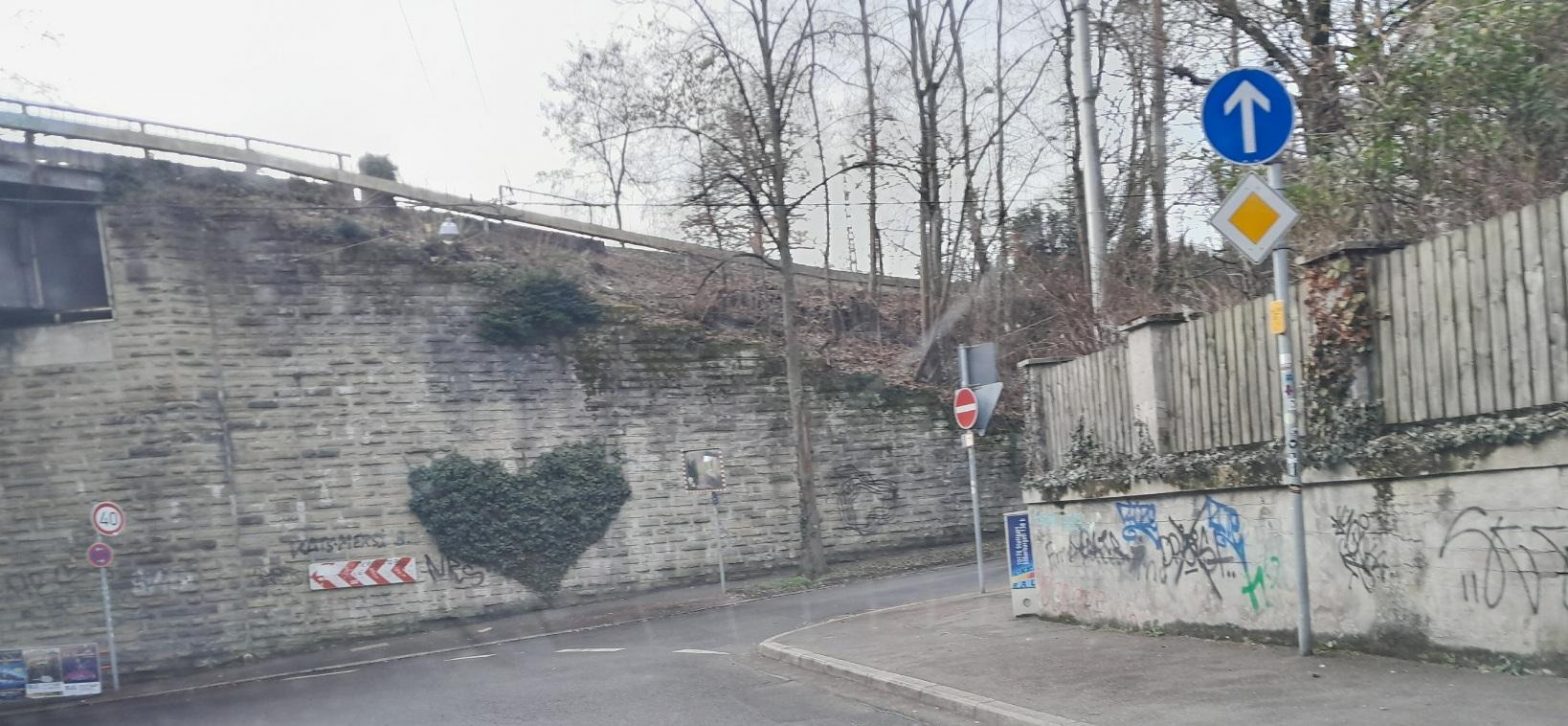 Eine Straßenecke mit einer Mauer, die zur Brücke über die Straße wird. An der Mauer eine grüne Kletterplfanze, die in Form eines Herzes geschnitten ist.